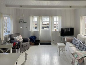 A living room with a sofa, a television, a dining table, and a window with curtains. - Thatched wooden house with sea view (Haderslev)