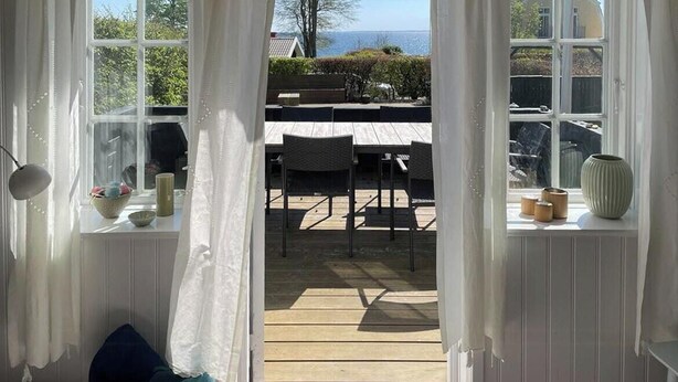 Ein Zimmer mit Meerblick durch ein Fenster, eine Holzterrasse davor und ein Stuhl sowie ein Tisch im Inneren.