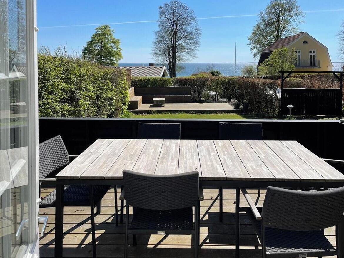 Un patio exterior con mesa y sillas de madera, un ventanal y vistas a una casa y árboles.