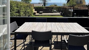 Eine Terrasse im Freien mit einem Holztisch und -stühlen, einer Glastür und Blick auf ein Haus und Bäume.