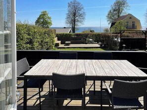 An outdoor patio with a wooden table and chairs, a glass door, and a view of a house and trees. - Thatched wooden house with sea view (Haderslev)