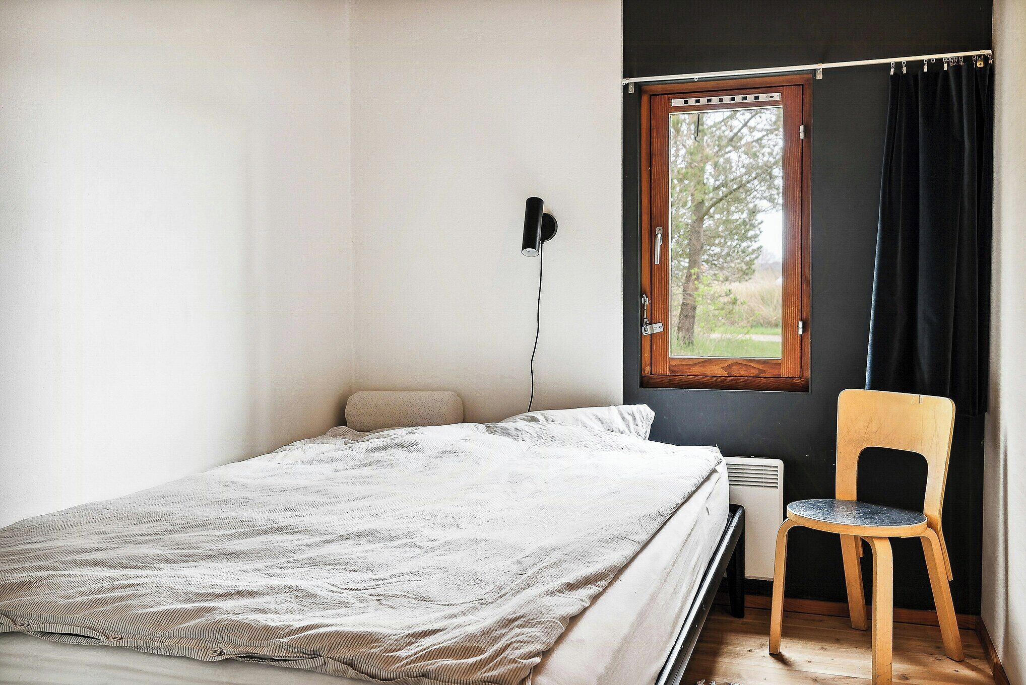 Een slaapkamer met een bed, een stoel, een raam met gordijnen en een wandlamp.