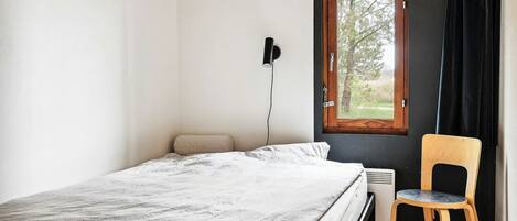Een slaapkamer met een bed, een stoel, een raam met gordijnen en een wandlamp.