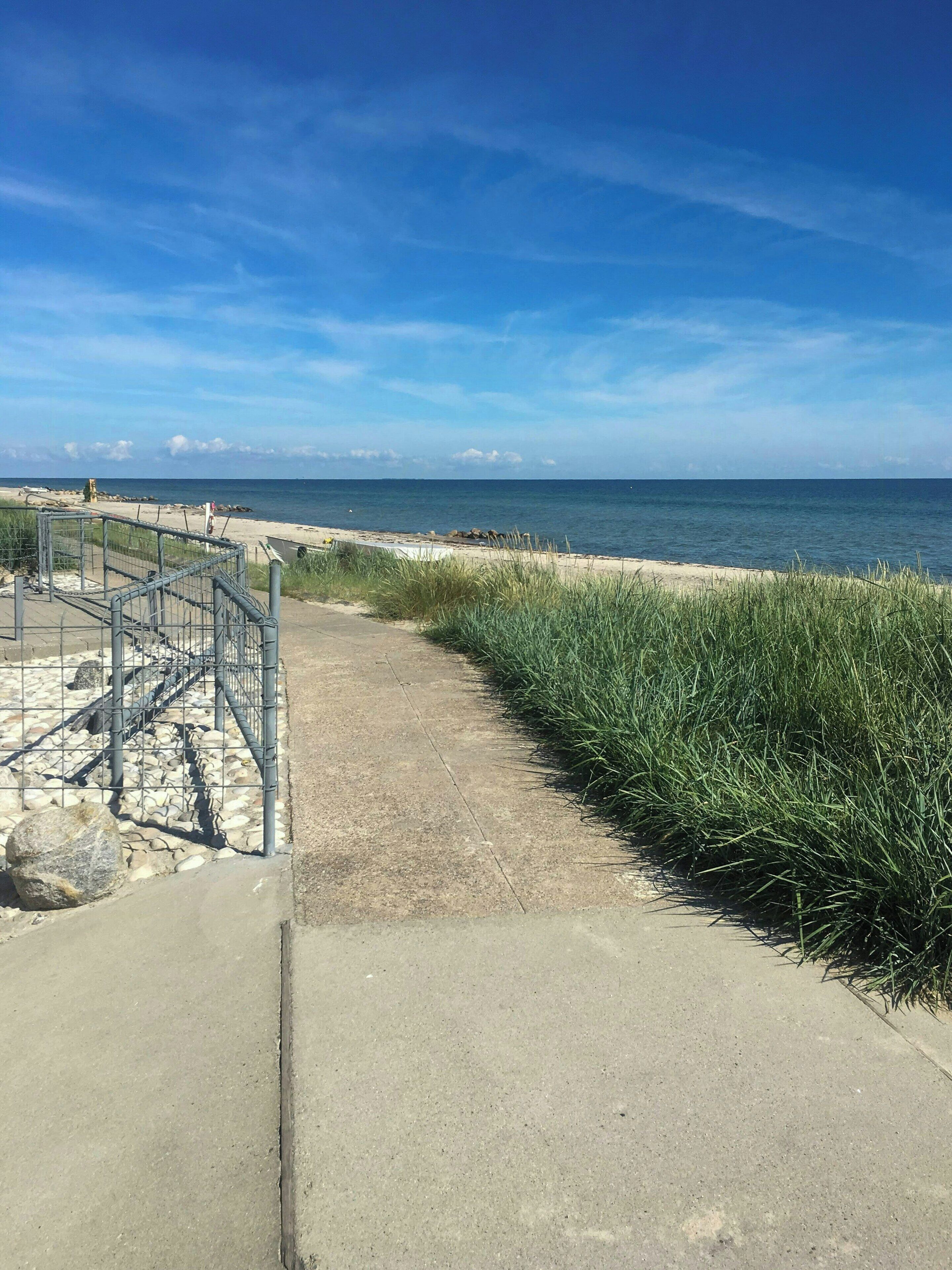 A concrete pathway leading to a sandy beach with a metal fence on the left and tall grass on the right.