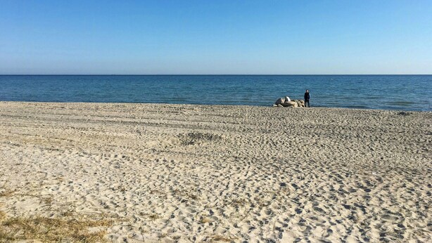 Ein Strand mit blauem Himmel, eine Person steht in der Nähe von Felsen, und im Hintergrund das Meer.
