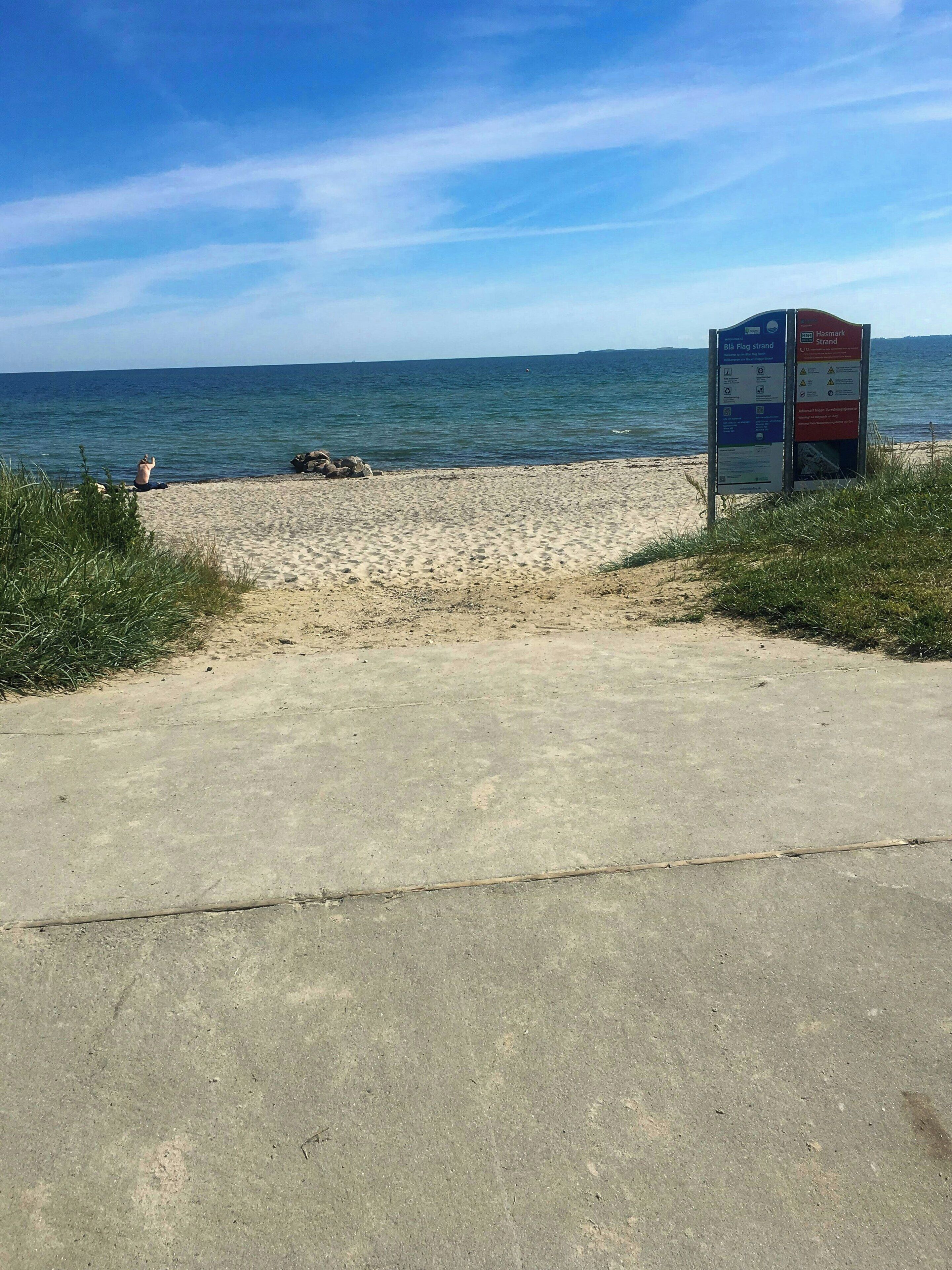Une plage avec une promenade en béton, un panneau indicateur et l’océan en arrière-plan.