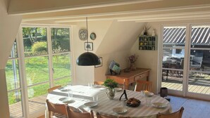 Un coin repas avec une table dressée pour quatre personnes, entourée de chaises en bois, d’un luminaire suspendu et offrant une vue sur l’extérieur grâce à de grandes fenêtres.