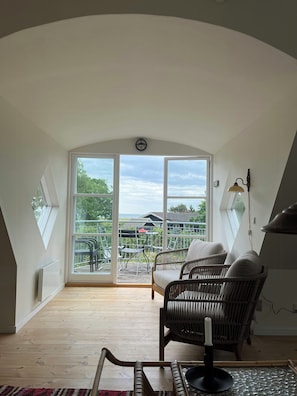 Ein Zimmer mit schrägem Dach, großen Fenstern, Korbmöbeln und Blick ins Grüne.