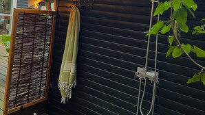 Un mur extérieur en bois avec un volet noir, un banc en bois sur lequel sont posées des bouteilles, une serviette suspendue et une pomme de douche.