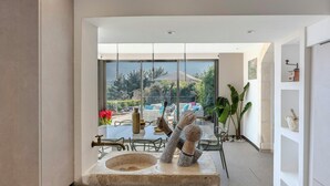 Um interior moderno com uma pia de pedra, área de jantar com cadeiras e mesa, e vista para um jardim e piscina através de grandes janelas.