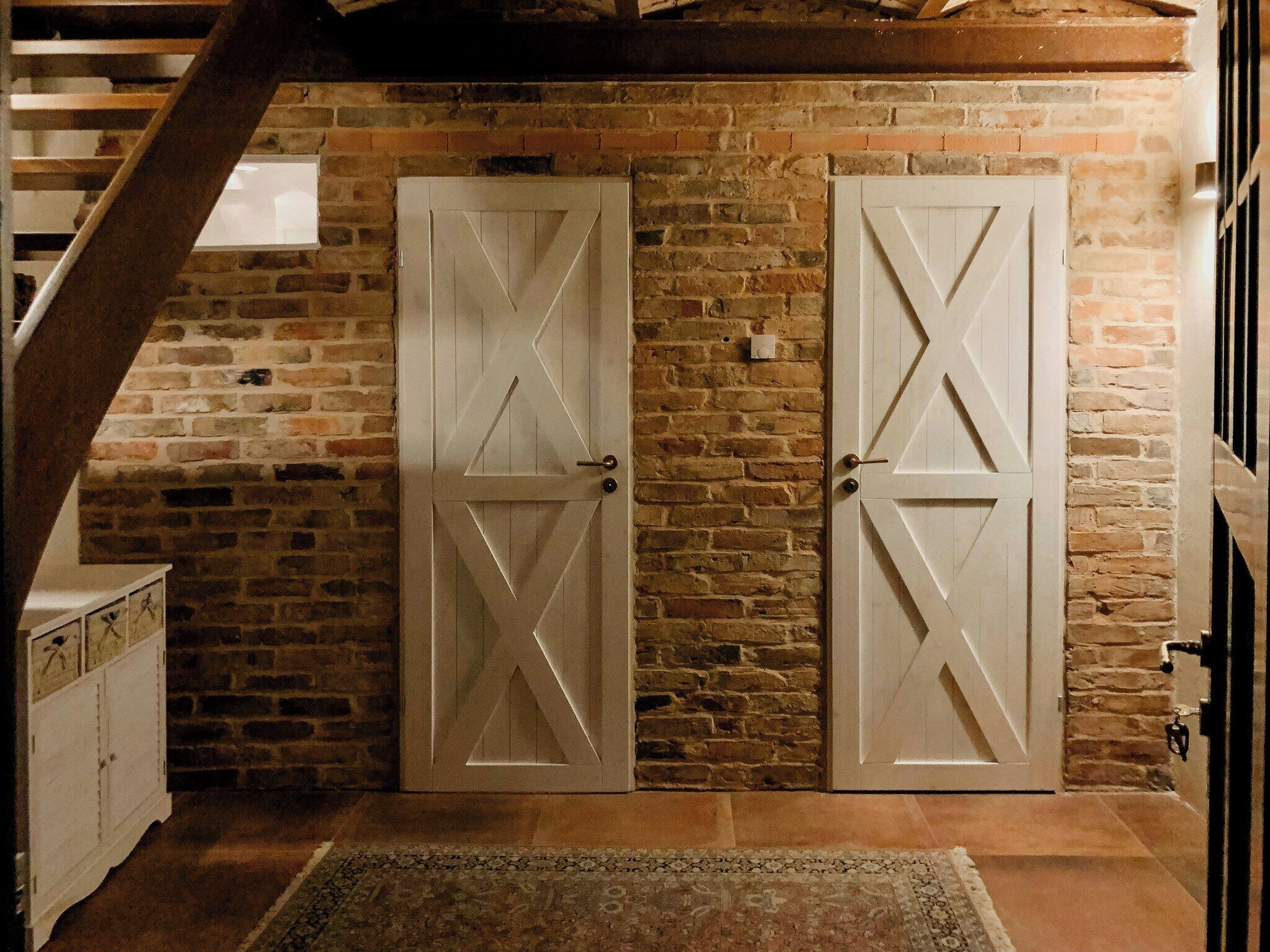 A room with two white doors with geometric patterns, a brick wall, a wooden staircase, and a patterned rug.