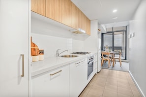 A modern kitchen with white cabinets, a stainless steel sink, wooden cabinets above the stove, and a dining area with a table and chairs. - 2BR CBD Escape-Pool, Gym & 17th floor views (Adelaide)