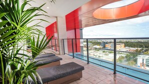 Ein Balkon mit Korbmöbeln, einer Glastrennwand und Blick auf eine Stadtlandschaft.