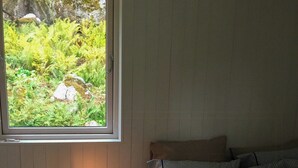 Um quarto com cama, mesa de cabeceira iluminada por um abajur, janela com vista para o verde e um tapete pequeno no chão.