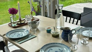 Mesa posta com pratos, copos e jarra, com vista para área verde lá fora.