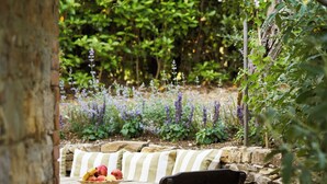Un mur de pierres, une table en bois avec un saladier de fruits et un coussin rayé.