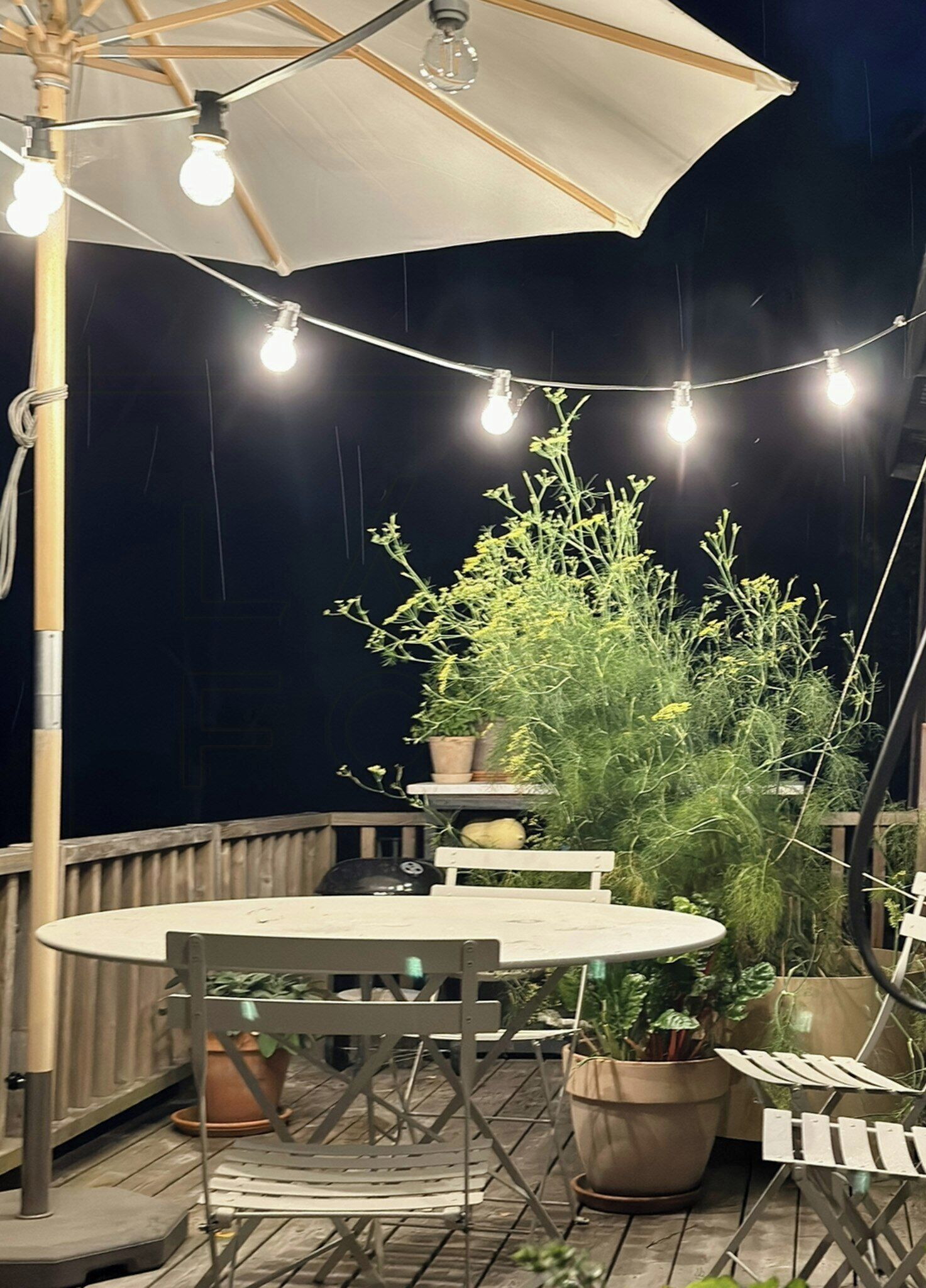 An outdoor patio with a round table, chairs, potted plants, string lights, and a large umbrella.