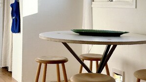 Een kamer met een ronde tafel, vier houten krukken, een raam met uitzicht op bomen, een hangmand en een blauwe tas aan de muur.