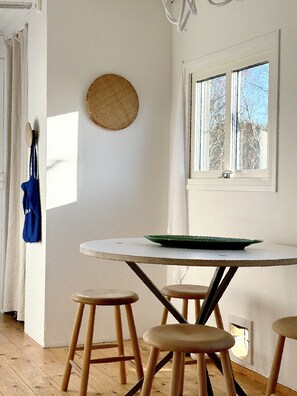 Ein Zimmer mit einem runden Tisch, vier hölzernen Hocker, einem Fenster mit Blick auf Bäume, einer hängenden Ampel und einer blauen Tasche an der Wand.