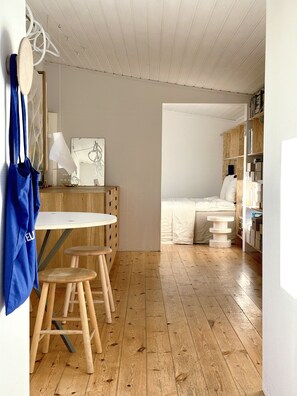 Ein kleiner Raum mit Holzboden, einem runden Tisch mit Hocker, einem Bett und einem Bücherregal.