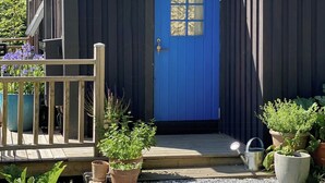Een klein, donker houten gebouw met een blauwe deur, omringd door potplanten en een grindpad.