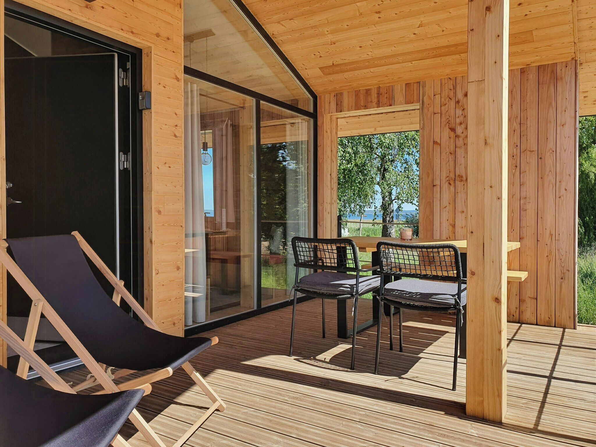 A wooden deck with two chairs and a table, a glass door leading inside, and a view of greenery outside.