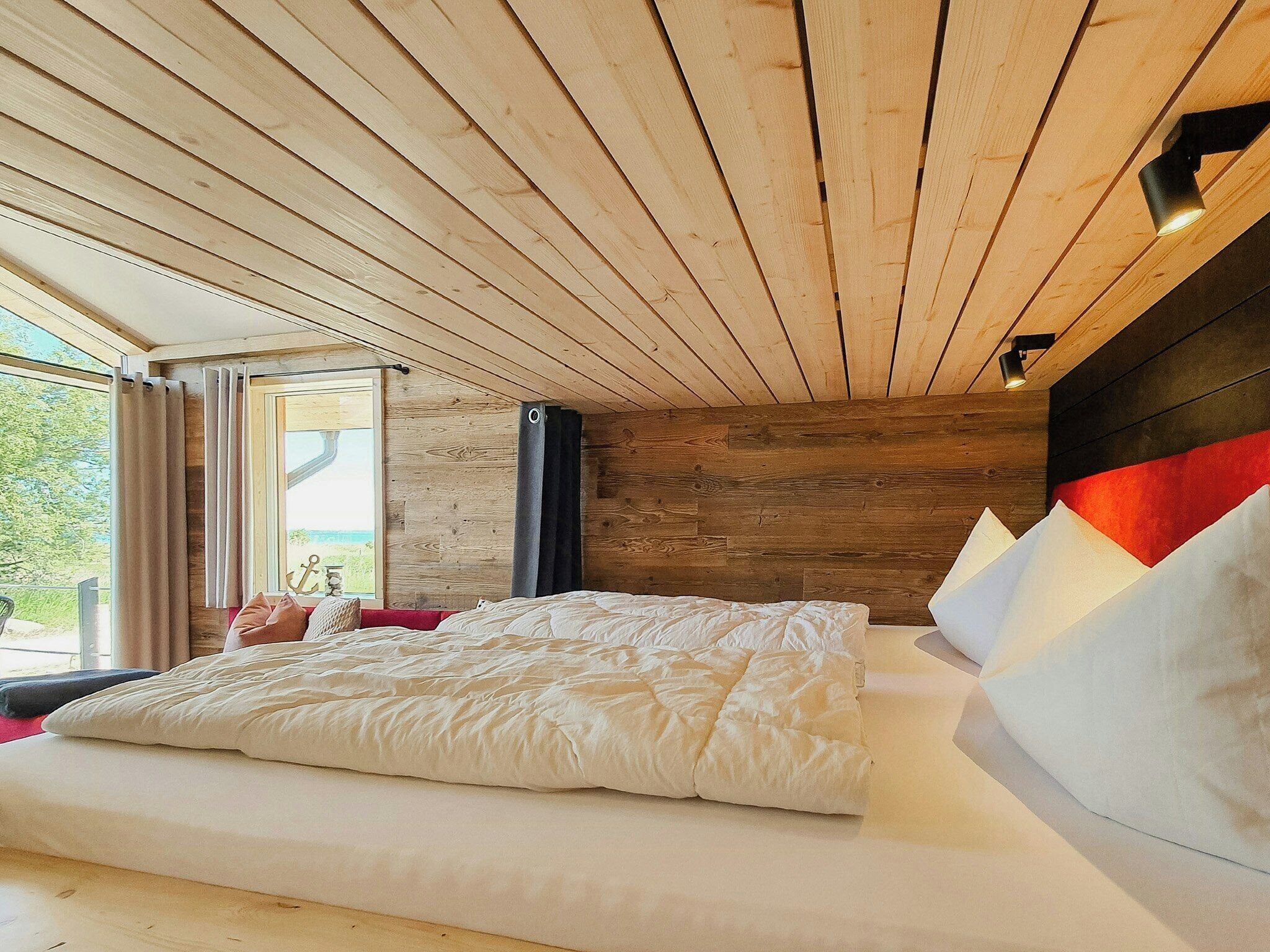 A bedroom with a wooden ceiling, a large bed with white bedding, a window with curtains, and a view of greenery outside.