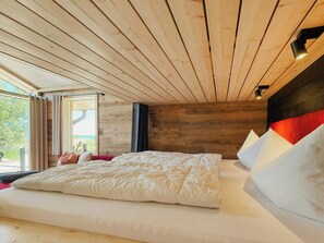 Ein Schlafzimmer mit Holzbalkendecke, einem großen Bett mit weißer Bettwäsche, einem Fenster mit Vorhängen und Blick auf Grünfläche.