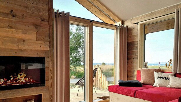 A room with a fireplace, a red sofa, and a large window with a view of the outdoors. - Tiny chalet with sea view at Werder (Boiensdorf)