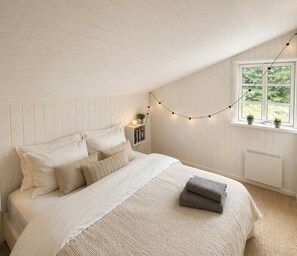 A bedroom with a sloped ceiling, a bed with white bedding, a bedside table with a lamp, a window with a view of greenery, and a string of lights. - Black wooden house in scenic surroundings (Blokhus)