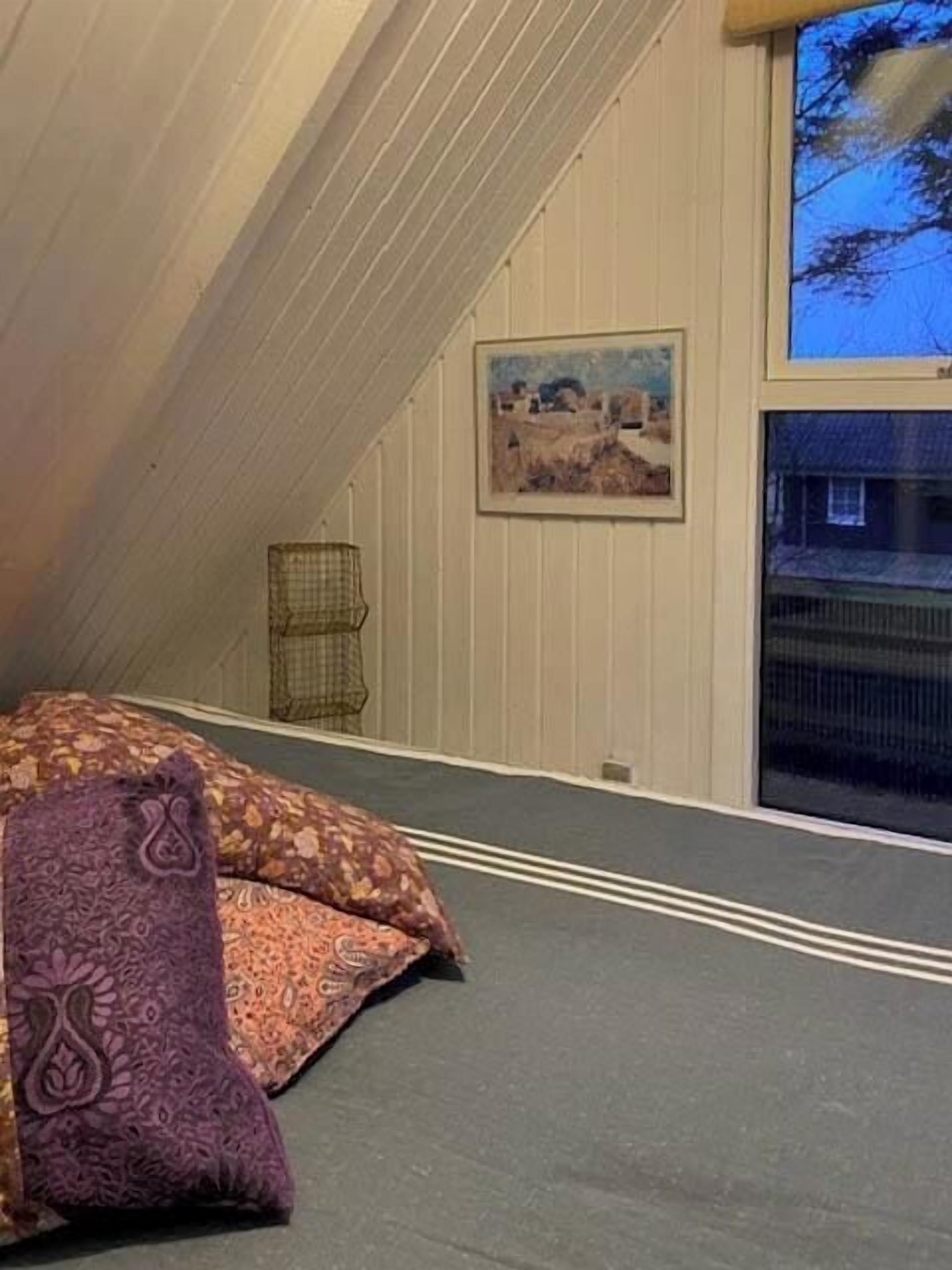 A bedroom with a sloped ceiling, a bed with patterned bedding, a framed picture on the wall, and a window with a view of a house.