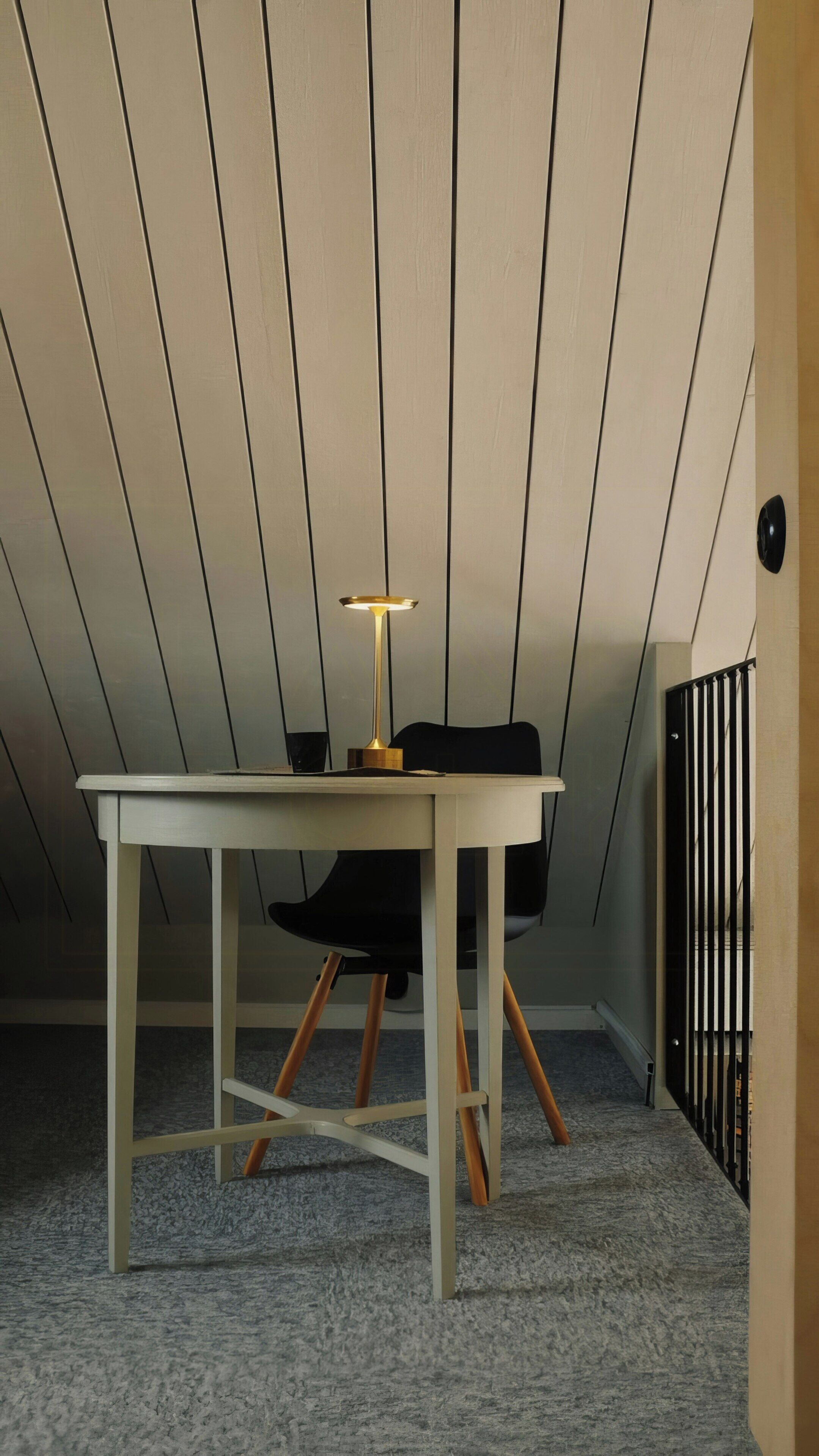Ein kleiner, gemütlicher Raum mit einem weißen Tisch und einem schwarzen Stuhl, eine Lampe auf dem Tisch und einer schrägen Decke.