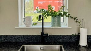 A kitchen with a stainless steel sink, a window with a view of a red barn, a potted plant, and a white paper towel holder.