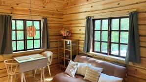 Interior de una cabaña de madera con un sofá, una mesa y dos ventanas con cortinas.
