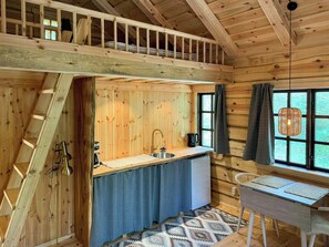 Ein Holzblockhaus-Interieur mit Schlafzimmer im Obergeschoss, einer Küche mit Spüle und Kaffeemaschine sowie einem Essbereich mit Tisch und Stühlen.