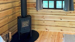 Interior de una cabaña de troncos con estufa de leña, una ventana con cortinas y una zona de estar acogedora.