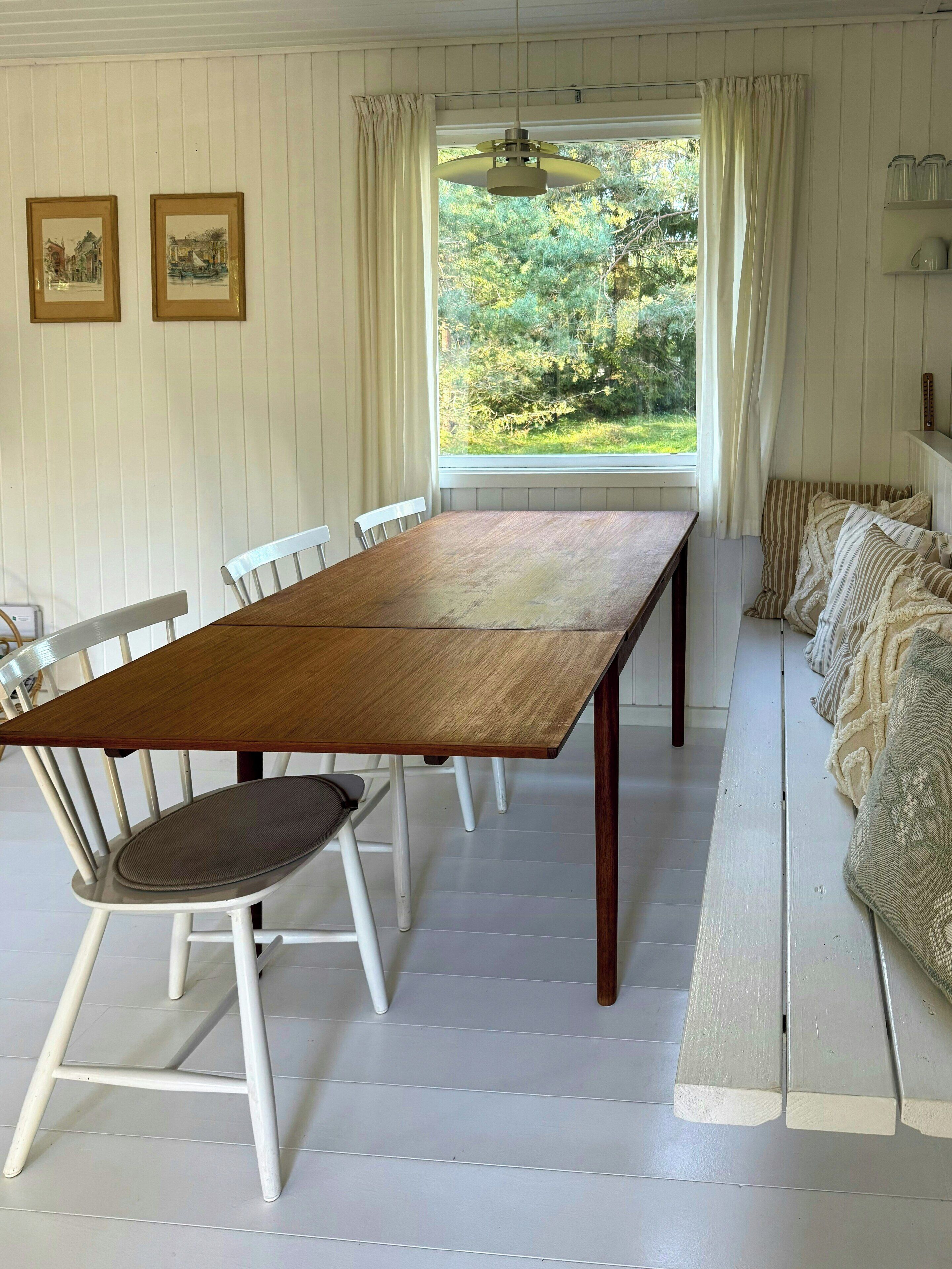 Un coin repas avec une table en bois, des chaises blanches, une vue par la fenêtre et un canapé agrémenté de coussins à motifs.