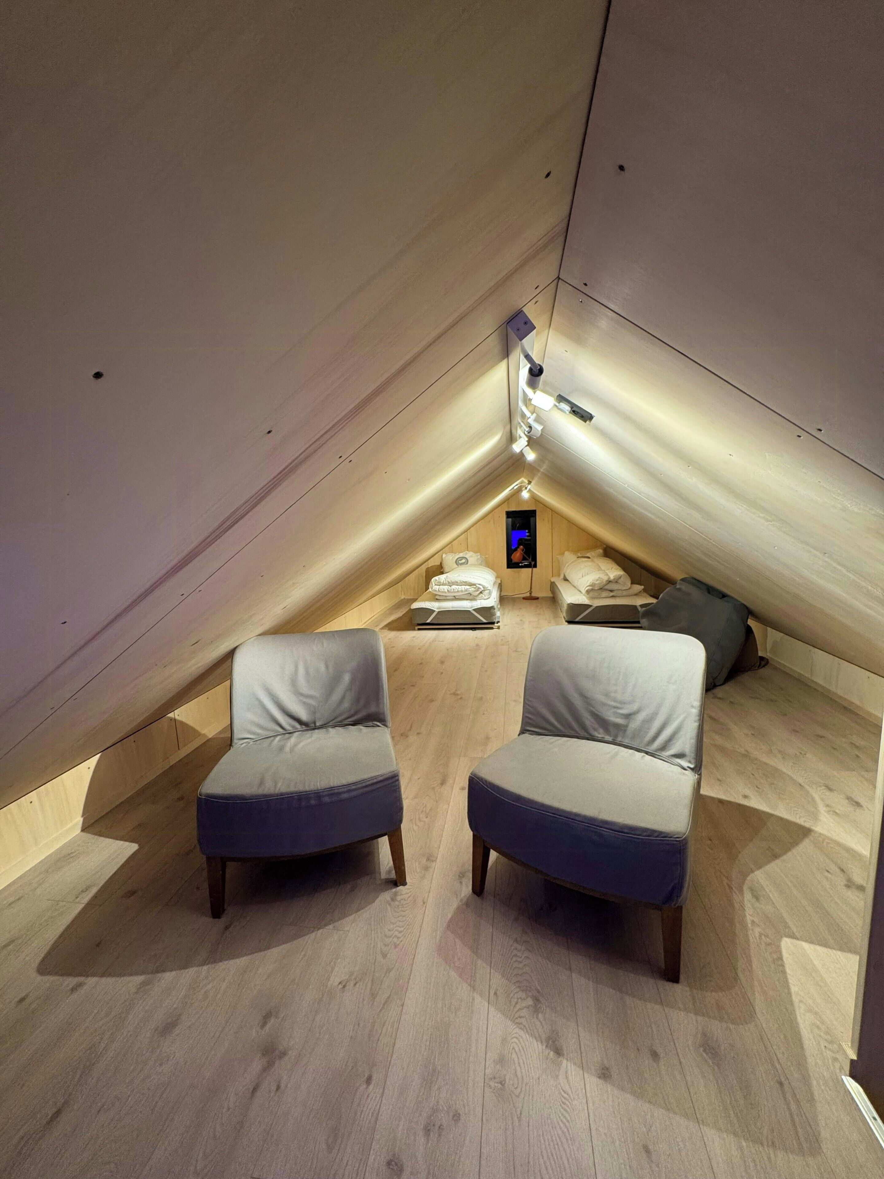 An attic bedroom with a sloped ceiling, two grey upholstered chairs, a small bed, and a built-in TV.