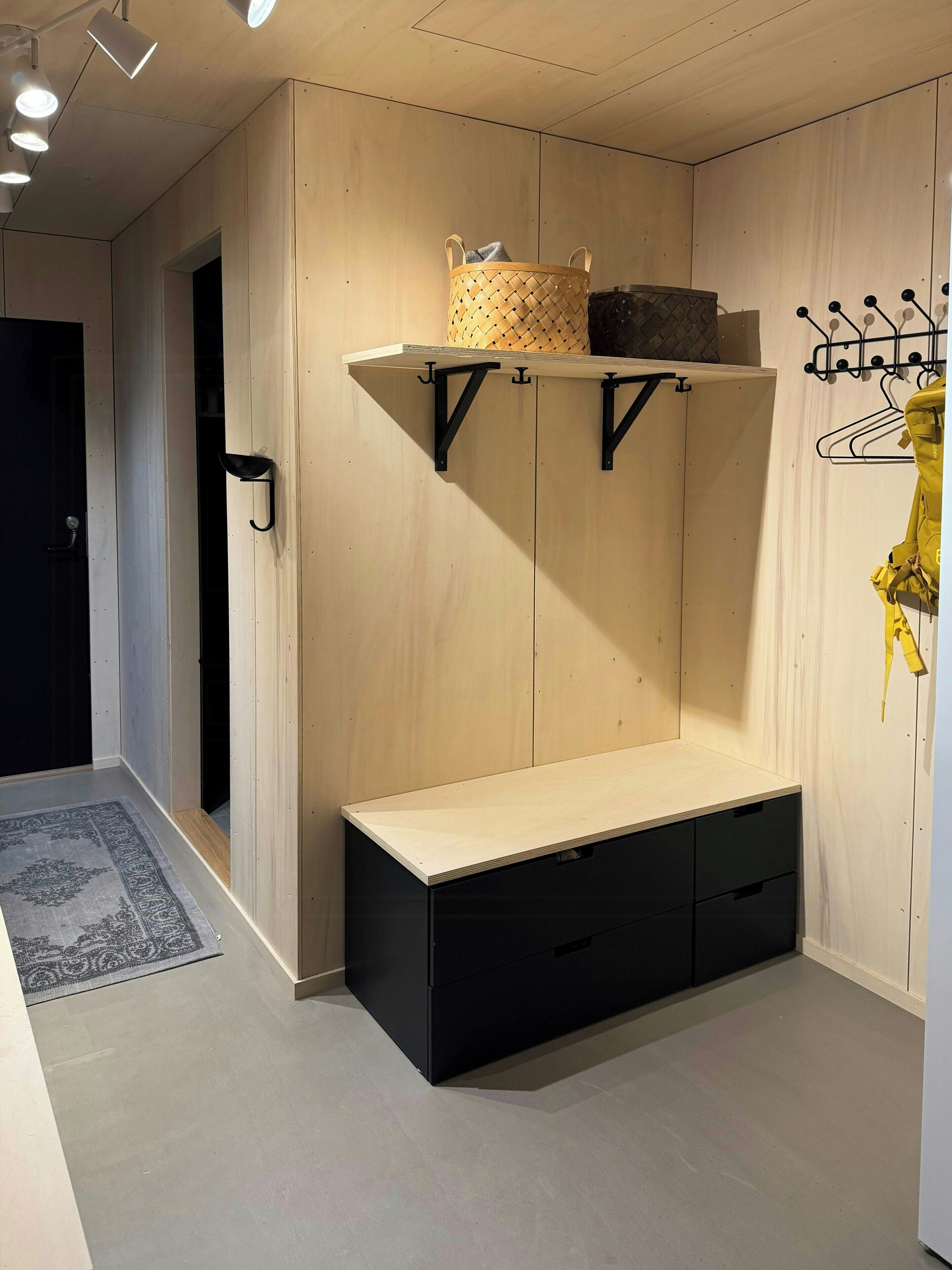A room with a wooden wardrobe, a bench with storage drawers, a hanging coat rack, and a patterned rug.