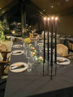 Una mesa de comedor preparada para una comida, con velas, copas y un árbol de Navidad al fondo.