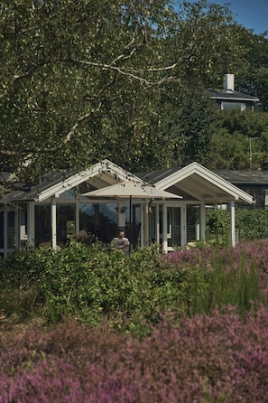 Ein Haus mit überdachter Veranda, umgeben von Grün und einem Hügel im Hintergrund.