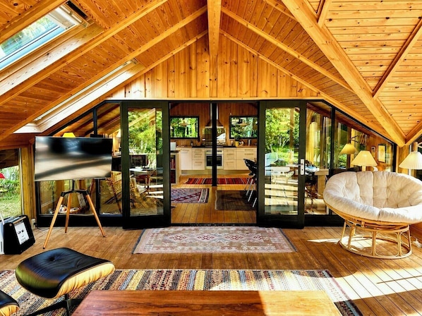 A spacious loft with a high ceiling, wooden beams, a dining area, a kitchen, and a large window offering a view of the outdoors. - Traditional Kalmar house close to the beach (sjaellands odde)