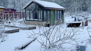 Un petit bâtiment enneigé, avec une porte et des fenêtres vertes, entouré d’arbres couverts de neige et d’une clôture.