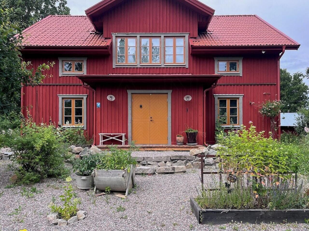 Una casa de madera roja con una puerta amarilla, dos ventanas en el piso superior y un camino de grava con plantas en macetas.