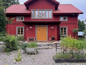 Una casa de madera roja con una puerta amarilla, dos ventanas en el piso superior y un camino de grava con plantas en macetas.