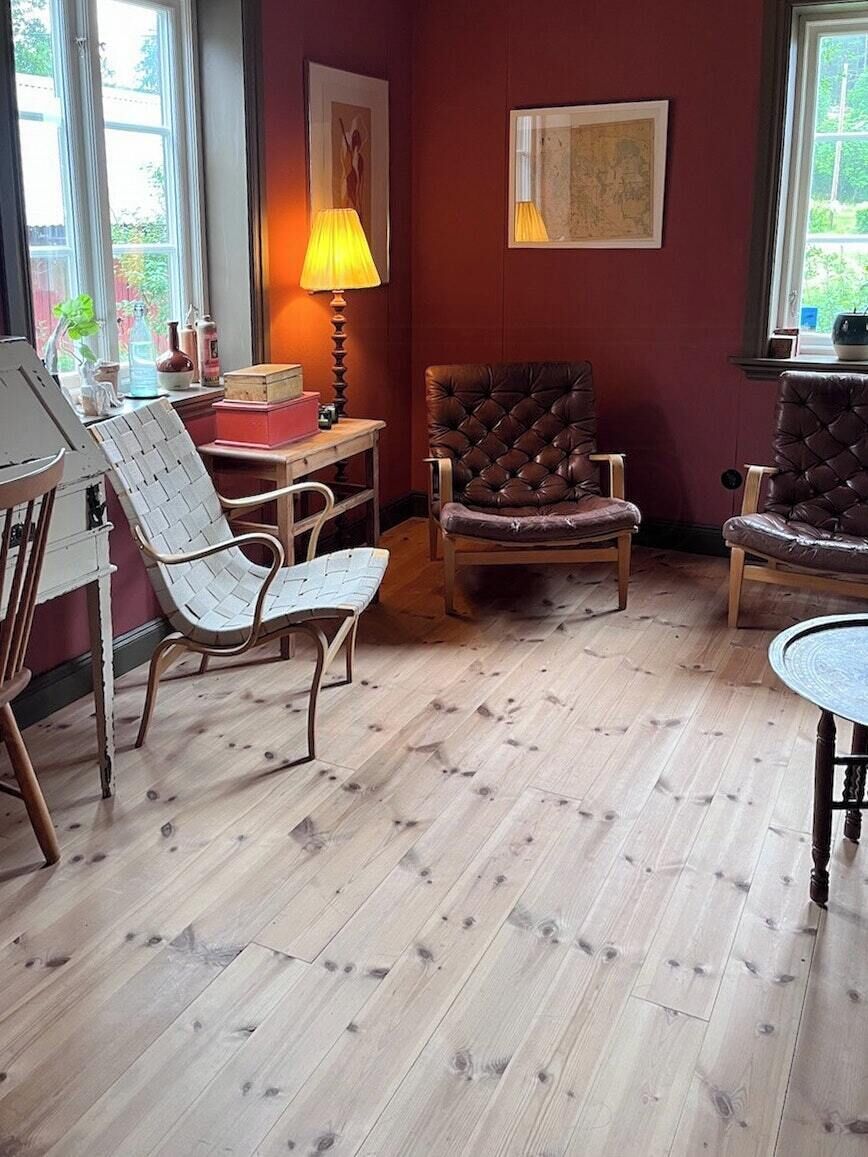 Una habitación con piso de madera, dos sillones de cuero, una silla blanca tejida, una mesita y una lámpara.
