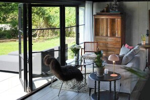 Ein Zimmer mit Glastür zu einem Garten, einem Holzschrank, einer Couch, einem kleinen Tisch mit Lampe und einem Stuhl.