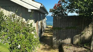 A narrow pathway leading to a house with a view of the ocean, surrounded by greenery and a wooden fence.