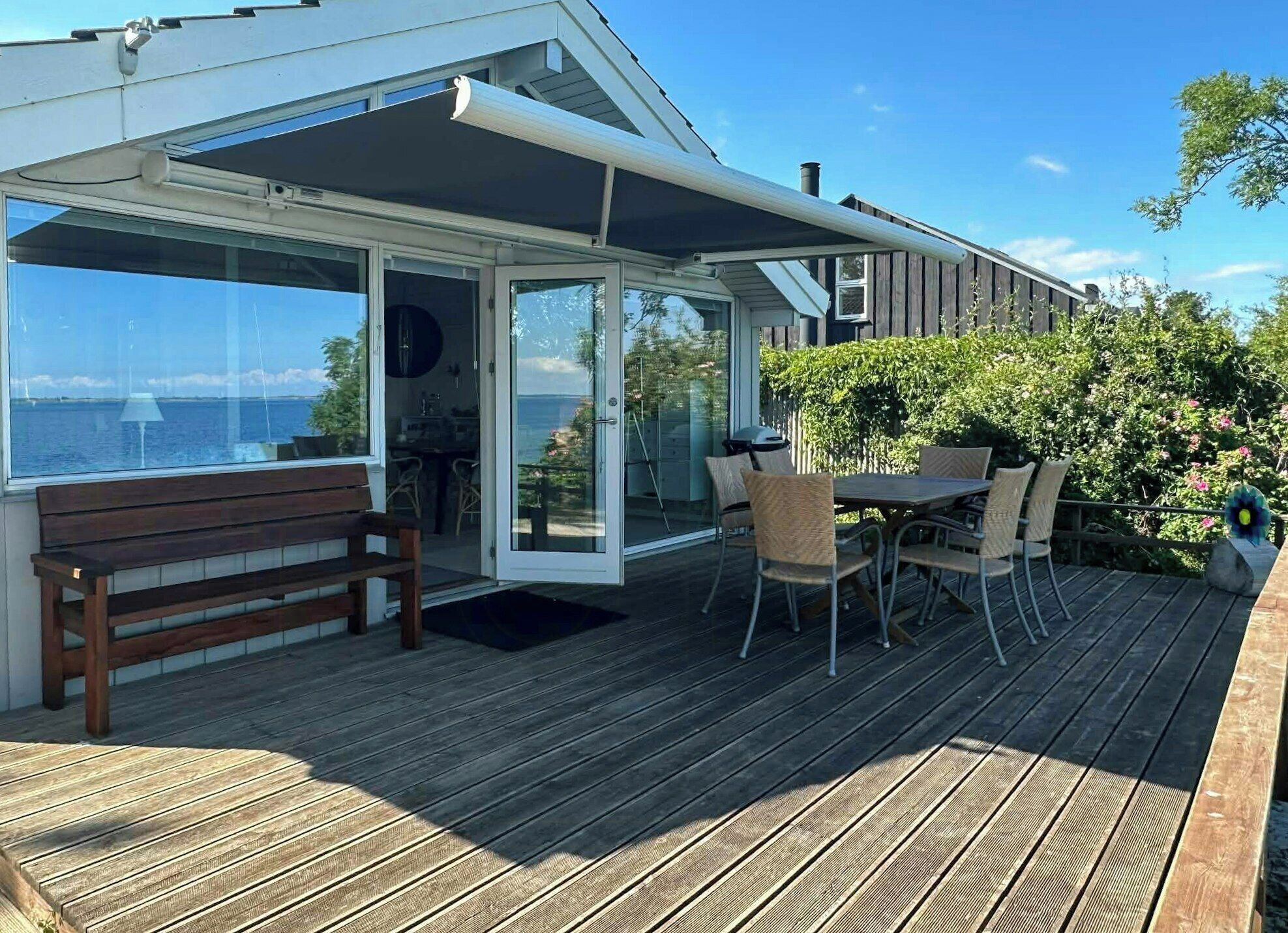 Una terraza de madera con una mesa y sillas, un banco y un ventanal de vidrio que da a un espacio interior.