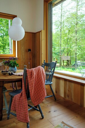 A wooden interior with a dining table, chairs, a vase with flowers, and a view of greenery outside.